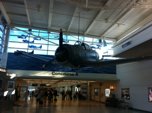 A vintage airplane hangs from the ceiling in an airport, with a mural of planes flying over water in the background.