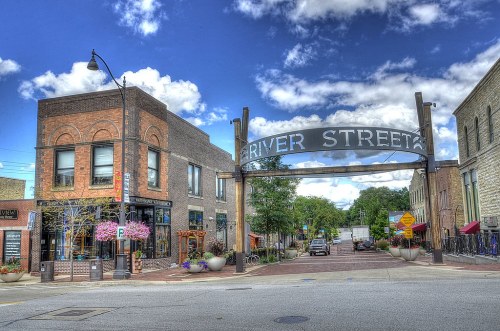 A vibrant street scene featuring shops and a large "River Street" sign under a blue sky with fluffy clouds.