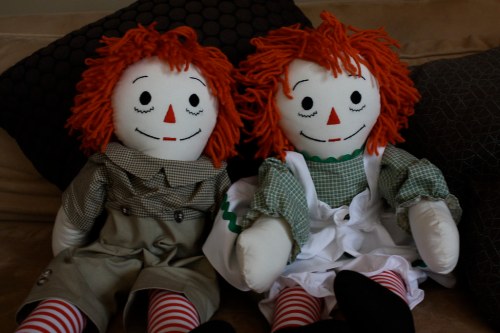 Two rag dolls with red yarn hair, smiling faces, and striped legs, sitting on a couch with patterned pillows.