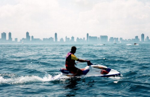 A person rides a jet ski on a calm water surface with a city skyline in the background under a cloudy sky.