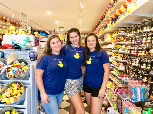 Three women in matching blue shirts pose together in a colorful store filled with toys and rubber ducks.