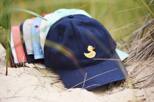 A navy cap with a yellow duck embroidery, surrounded by colorful caps on a sandy beach.