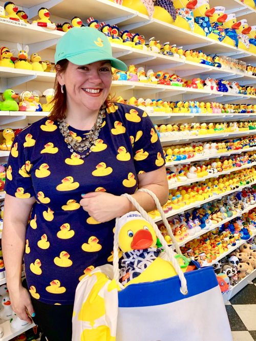 A smiling woman in a duck-patterned shirt and cap holds a bag with a large rubber duck, surrounded by shelves of ducks.