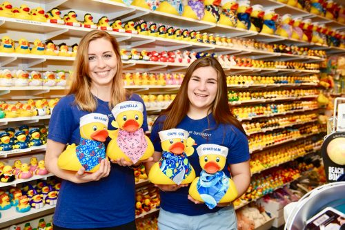 Two women hold colorful rubber ducks in a shop filled with rows of various rubber ducks in the background.