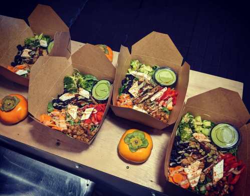 Four takeout boxes filled with colorful salads, vegetables, and a small container of dressing, arranged on a wooden table.
