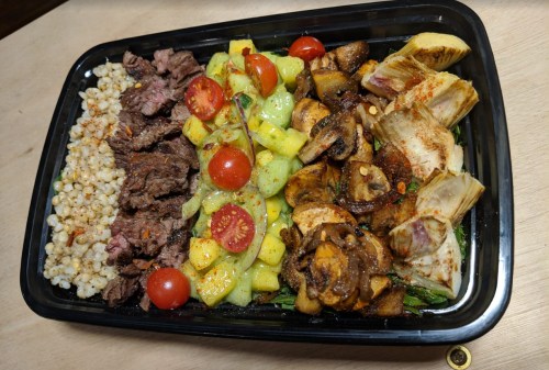 A black tray filled with grilled steak, cherry tomatoes, diced pineapple, mushrooms, and artichokes on a wooden surface.