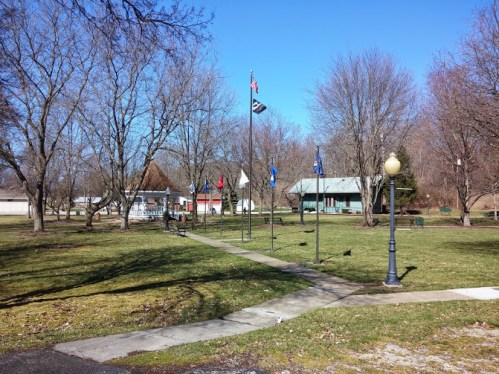 A park scene with bare trees, flags, a gazebo, and a pathway under a clear blue sky.