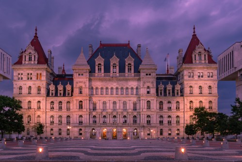 The New York State Capitol building illuminated at dusk, surrounded by trees and a plaza under a purple sky.