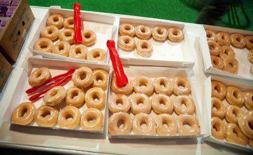 A display of glazed donuts in boxes, with red serving spoons on the side.