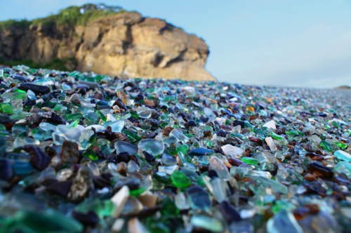 A close-up of a beach covered in colorful sea glass, with a rocky cliff in the background under a clear blue sky.