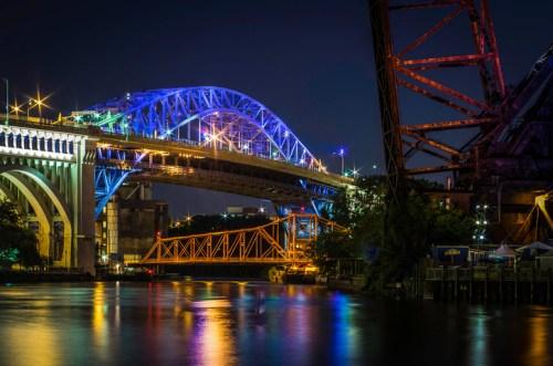 Night view of illuminated bridges over a river, with reflections in the water and city lights in the background.