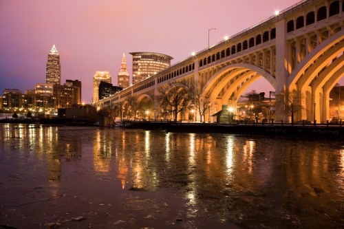 Cleveland skyline at dusk, featuring a bridge over icy water with city lights reflecting on the surface.