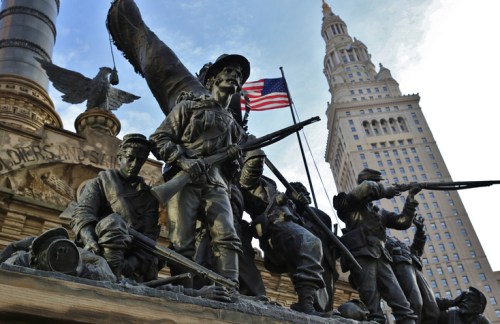 A bronze statue of soldiers with an American flag, set against a tall building and blue sky.