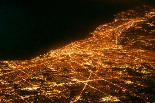 Aerial view of a city at night, showcasing a network of bright lights and streets against a dark background.
