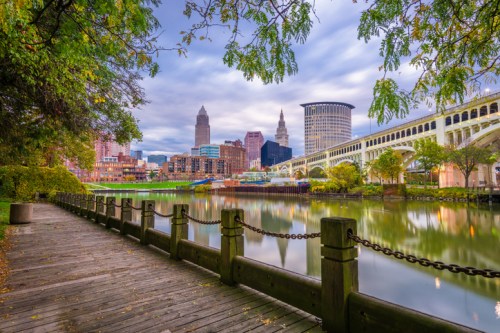 A scenic view of a city skyline reflected in a river, with trees and a wooden walkway in the foreground.