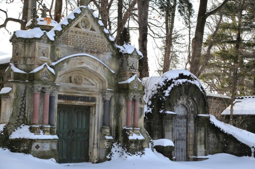 Snow-covered cemetery with ornate, weathered tombs surrounded by trees.
