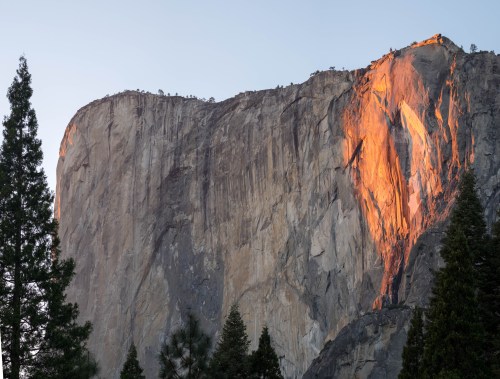 A dramatic cliff face illuminated by sunset, surrounded by evergreen trees in a serene landscape.