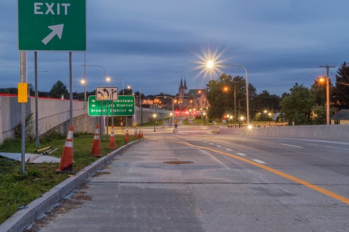 A road leading to an exit sign, with traffic lights and a church in the background under a twilight sky.