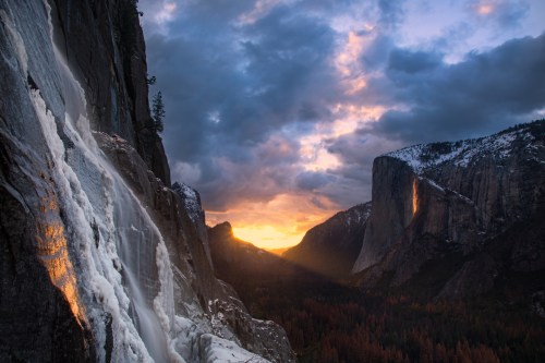 A stunning sunset over a valley, with a waterfall in the foreground and dramatic clouds above the mountains.