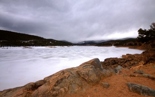 A serene winter landscape featuring a frozen lake surrounded by rocky shores and distant mountains under a cloudy sky.