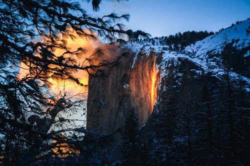 A stunning sunset illuminates a snowy mountain, casting a warm glow on the rocky cliff amidst silhouetted trees.