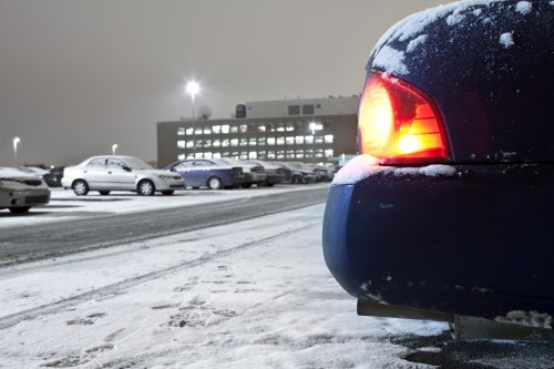 A snowy parking lot at night, featuring a blue car with its brake light on and a building illuminated in the background.