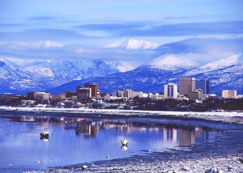 A snowy landscape with a city skyline reflecting in water, surrounded by mountains under a blue sky.