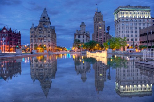 A cityscape at dusk featuring historic buildings reflected in a calm waterway.