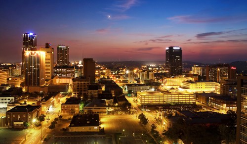 A city skyline at dusk, with illuminated buildings and a colorful sky transitioning from day to night.