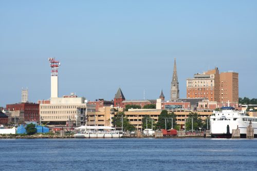 City skyline featuring buildings, a church steeple, and a waterfront with boats under a clear blue sky.