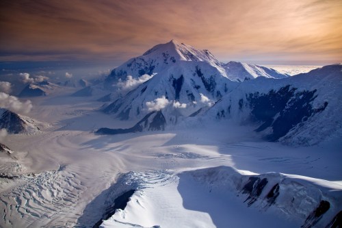 Aerial view of a snow-covered mountain range with glaciers and clouds under a colorful sky at sunset.