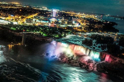 Aerial view of Niagara Falls illuminated at night, with colorful lights and a city skyline in the background.