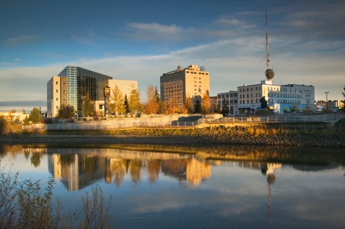 Cityscape reflecting in water, featuring modern buildings and trees under a clear blue sky.