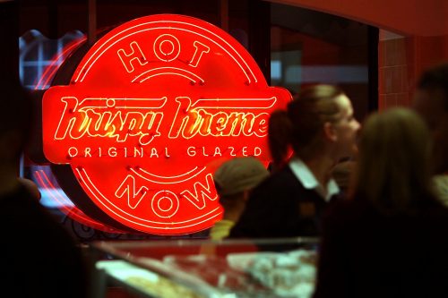 Neon "Hot Now" sign at a Krispy Kreme shop, with blurred customers in the foreground.