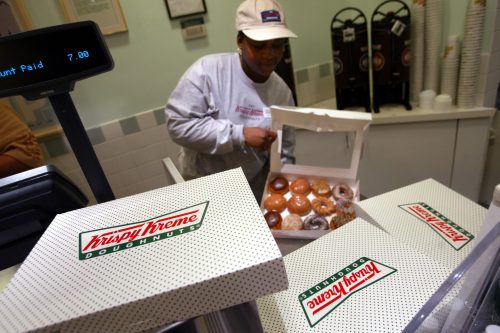 A woman at a counter holds a box of Krispy Kreme donuts, with additional boxes displayed nearby.