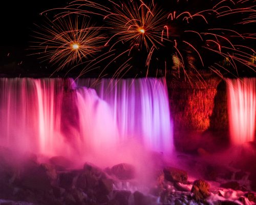 Colorful fireworks illuminate a waterfall at night, with vibrant lights reflecting off the water's mist.