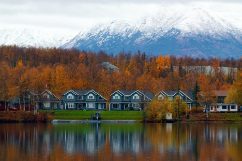 A serene lake reflects colorful autumn trees and houses, with snow-capped mountains in the background.