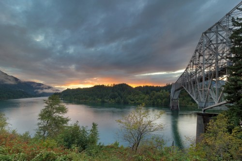 A scenic view of a bridge over a calm river, surrounded by lush greenery and a dramatic cloudy sky at sunset.