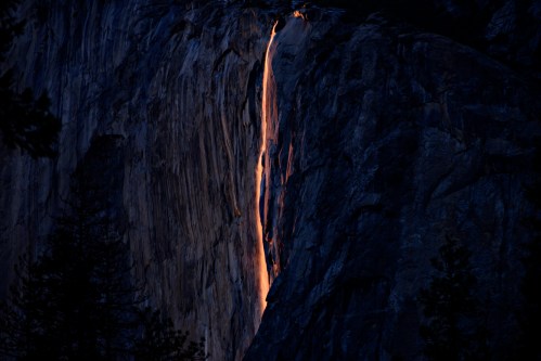 A glowing waterfall cascades down a dark rock face, illuminated by sunset, creating a striking natural spectacle.