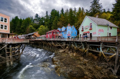 Colorful wooden buildings on stilts by a flowing river, surrounded by lush green trees under a cloudy sky.