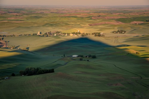 Aerial view of rolling green hills with shadows, dotted with small farms and trees under a soft evening light.