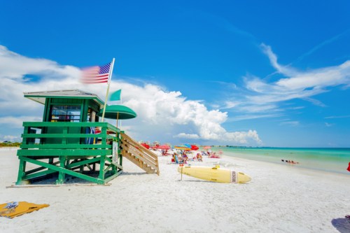 A colorful lifeguard tower on a sunny beach, with umbrellas, chairs, and calm waves under a blue sky.