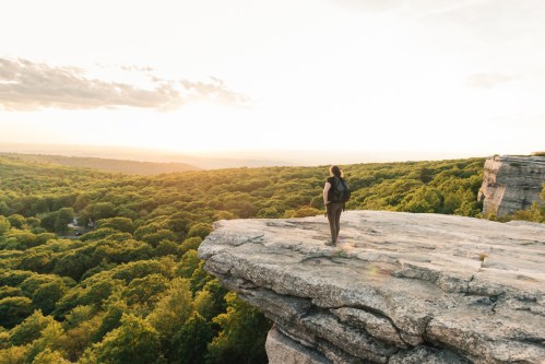 A person stands on a rocky ledge overlooking a lush green forest at sunset.