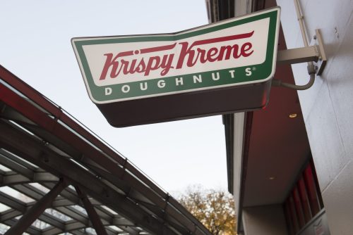 Sign for Krispy Kreme Doughnuts hanging outside a store, with a clear sky in the background.