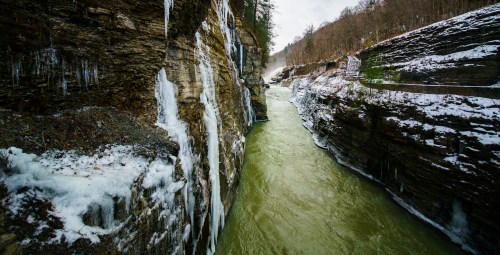A narrow river flows between steep, icy cliffs, surrounded by winter scenery and misty trees.
