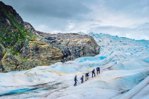 A group of climbers traverses a blue glacier surrounded by rocky terrain and cloudy skies.