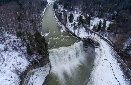 Aerial view of a waterfall cascading into a river, surrounded by snowy trees and a winding path.