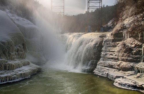 A frozen waterfall cascades over rocky cliffs, with mist rising and a bridge in the background.