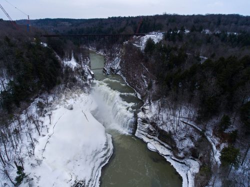 Aerial view of a snowy waterfall cascading into a river, surrounded by trees and a winter landscape.