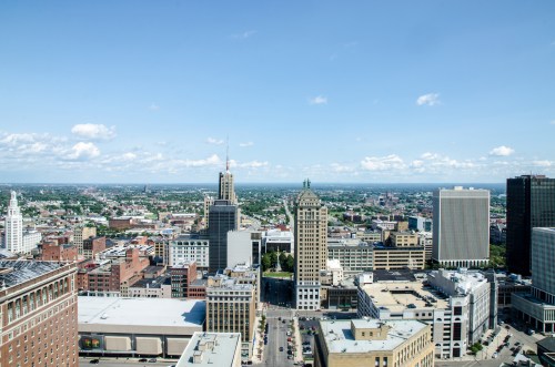 A panoramic view of a city skyline with various buildings under a clear blue sky and scattered clouds.